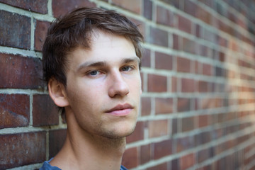 Portrait of a young man leaning against a brick wall with a skeptic and serious expression on his face.