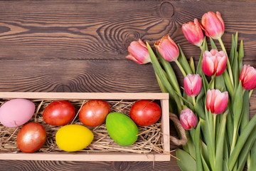 Top view of easter colorful eggs on a bed of straw in a long wooden box on a wooden table and red tulips, space for text.