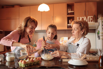 Grandmother and mother teaching teenage girl how to make muffin dough, mixing ingredients with wire whisk in their home kitchen