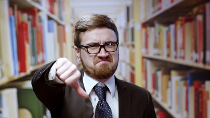 Dissatisfied young caucasian businessman in suit showing negative sign thumbs down of disregard looking upset posing near bookshelves in the library.