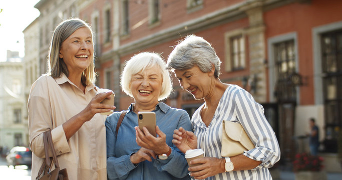 Three beautiful and cheerful Caucasian women with coffee to-go in hands laughing while scrolling photos on smartphone outdoor. Happy joyful grandmothers smiling while watching pictures on smartphone.