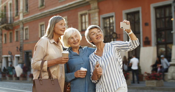 Three Caucasian beautiful old women smiling while taking selfie photo with smartphone camera on street in city center. Senior happy ladies posing to phone while taking selfies picture.
