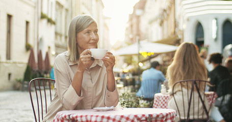 Good looking senior Caucasian smiled woman with gray hair sipping coffee while sitting at table in cafe terrace outdoors. Lady on retirement drinking tea and resting. Pensioner spending time nice.