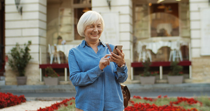 Caucasian Beautiful Old Woman Standing In Center City And Tapping Message On Smartphone. Happy Grandmother Using Cellphone While Typing Sms. Senior Nice Female Texting And Scrolling On Mobile Phone.