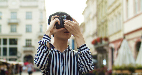 Close up of handsome Asian young man traveller with backpack taking photo with photocamera in center city of european town. Chinese guy student tourist having sightseeing tour. Traveling concept.