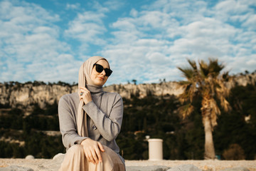 Fototapeta premium portrait of young European Muslim women with hijab sitting on the stone beach with sea in the background. She is happy and relaxed.