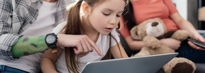 Panoramic shot of woman using laptop with daughter near parent with teddy bear on couch