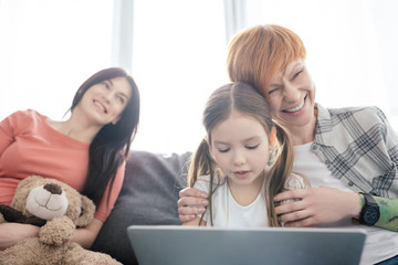 Selective focus of cheerful mother hugging daughter with laptop near smiling parent with teddy bear...