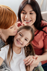 Cheerful kid smiling at camera near same sex parents on couch