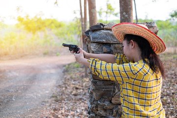 Fototapeta premium Portrait the farmer asea woman wearing a hat at the shooting shot from old revolver gun in the farm, Young girl sitting in the attitude of aiming and looking through the sight handgun