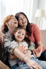 Selective focus of same sex parents laughing while embracing daughter on couch in living room