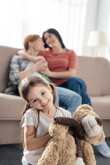 Selective focus of child with teddy bear smiling at camera near same sex parents embracing on couch...