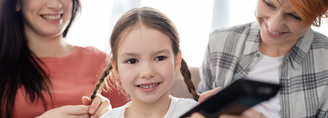 Panoramic shot of smiling mothers sitting near cheerful child with remote controller on couch