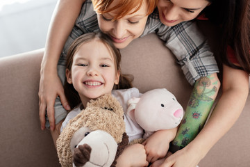 Smiling mothers hugging happy child with soft toys looking at camera on couch