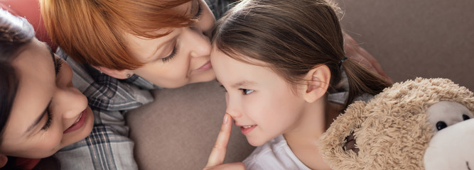 Panoramic shot of smiling same sex parents kissing and touching nose of daughter with teddy bear on...