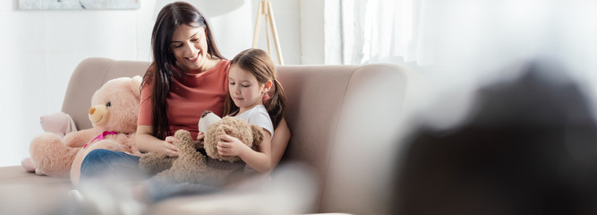 Selective focus of kid playing with teddy bear near smiling mother on sofa in living room,...