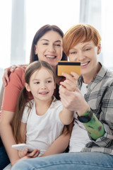 Selective focus of smiling kid with smartphone holding credit card near same sex parents on sofa
