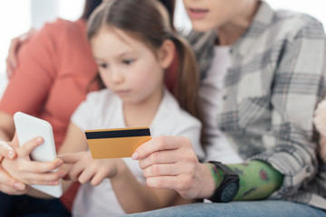 Selective focus of same sex parents holding credit card while daughter using smartphone at home