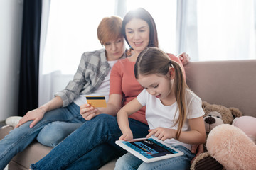 Kid using digital tablet with booking app on screen near mothers with credit card at home