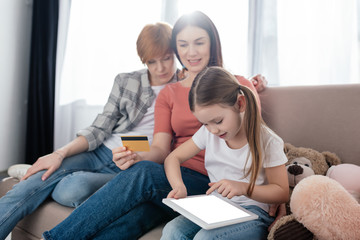 Same sex couple with credit card sitting near daughter with digital tablet on couch