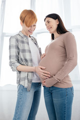 Smiling woman touching belly of pregnant girlfriend at home