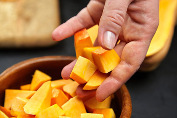 Pumpkin pieces in female hand. Chopped butternut squash in a wooden bowl on black background,...