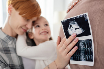 Selective focus of mother hugging smiling daughter and holding ultrasound scan of baby near belly...