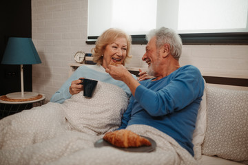 Laughting elderly couple is having good time while sitting on bed in bedroom. Elderly men offering his wife with croissant while she drinks morning coffee.