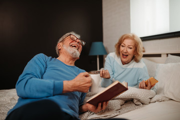 Laughting senior couple lying on bed. Elderly man with classes is holding cup of coffee and reading book while his wife lying next to him and holding smartphone.
