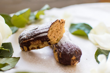 French eclairs with custard sprinkled with powdered sugar and white roses, petals, green leaves on light background
