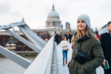 Young pretty hipster woman wearing winter hat, coat and trendy leather waist bag on Millennium bridge in London relaxing, taking a break, looking straight