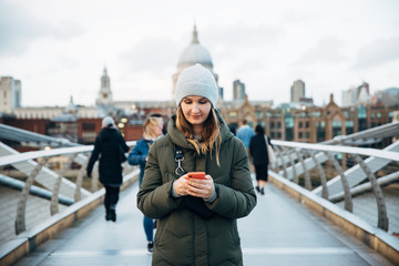 Attractive hipster woman using smartphone on millennium bridge in London on winter cloudy day. Travel app concept.