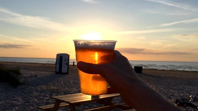 Glass of beer on sunset beach and man with beer and chips on background. Street cafe, wooden  table, male hand, plastic glass