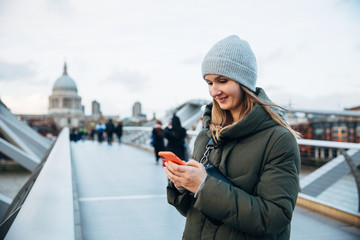 Attractive smiling woman in hat and coat using mobile phone travel application on Millennium bridge in London on cloudy winter day