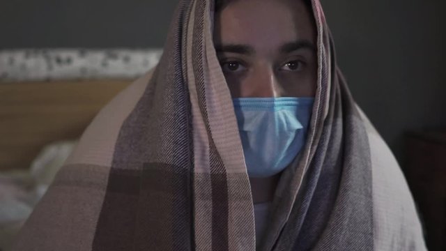 A sick man in the medical mask sitting under a blanket at home on quarantine