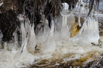 Ice sculptures created by frost and fallen trees in streaming river. Winter wonderland in Estonia. Icy stalagmites between branches and water surface. Cold winter day.