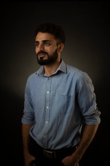 Fashion portrait of an young brunette Indian/European/Arabian/Kashmiri man in blue formal shirt and trousers in front of a black copy space background. Indian lifestyle and fashion photography.