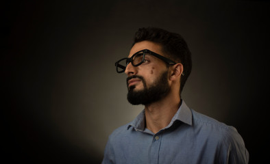 Portrait of young brunette Indian/European/Arabian/Kashmiri man in blue formal shirt and trousers with glasses in front of black copy space background. Indian lifestyle and fashion photography.