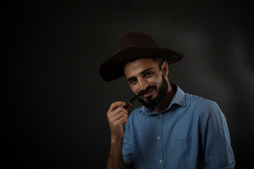 Portrait of young brunette Indian/European/Arabian/Kashmiri man in blue formal shirt and trousers with cowboy hat and pipe in front of black copy space background. Lifestyle and fashion photography.