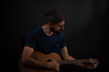 Portrait of a Kashmiri/European/Arabian man in casual wear and head band with playing guitar in front of a black copy space studio background. Indian lifestyle and fashion photography.