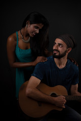 A dark skinned Indian/African girl and a Kashmiri/European/Arabian man in casual wear with guitar in front of a black copy space studio background. Indian lifestyle and fashion photography.