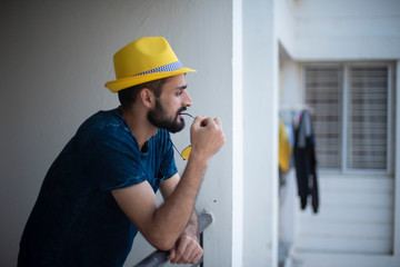 Portrait of young brunette Indian/European/Arabian/Kashmiri man in casual tee shirt and yellow hat with spectacles standing on a balcony in white background. Indian lifestyle.