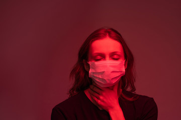 Infected young woman in face mask keeping eyes closed and feeling pain in throat, dark red light effect
