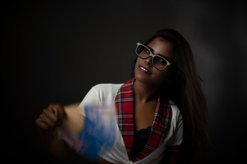 Portrait of an Asian/Indian/African brunette dark skinned young girl in sexy school uniform and spectacles with a Chinese fan in a black/grey studio background. Fashion and cosplay photography.