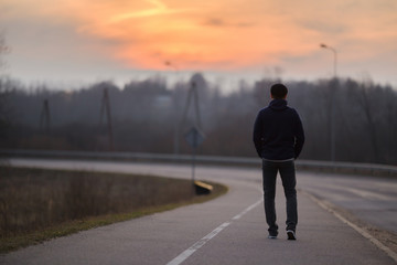 Young man alone slowly walking on sidewalk in dark evening. Orange sunset light in sky. Back view.