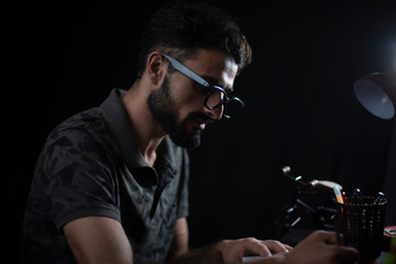 Portrait of young brunette Indian/European/Arabian/Kashmiri man in casual tee shirt with glasses studying in front of a table lamp in black copy space studio background. Indian lifestyle.
