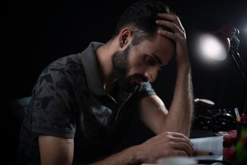 Fototapeta na wymiar Portrait of young brunette Indian/European/Arabian/Kashmiri man in casual tee shirt studying in front of a table lamp in black copy space studio background. Indian lifestyle.