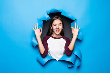 Young pretty woman with okay gesture while looking through blue hole in paper wall.