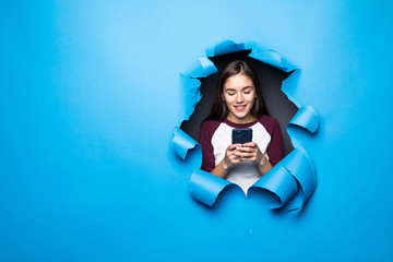 Young pretty woman typing and use phone while looking through blue hole in paper wall.