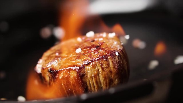 Filet mignon steak is fried in a pan with fire. Salt falls on a steak, slow motion.